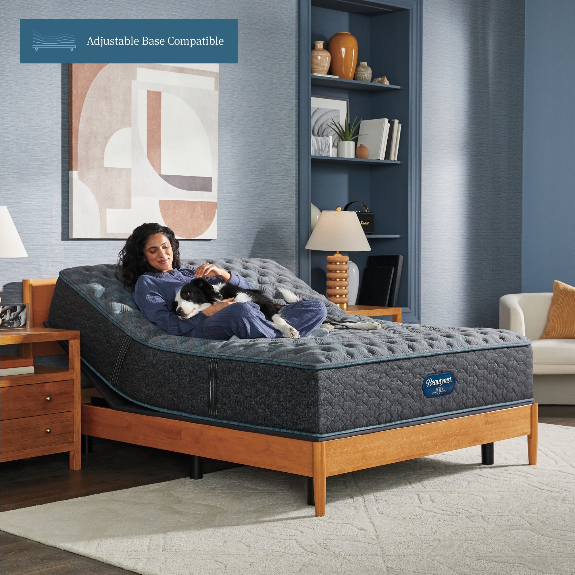 Woman sitting on a slightly elevated on an adjustable base Beautyrest Innerspring Mattress sitting in a brown bed frame holding her dog